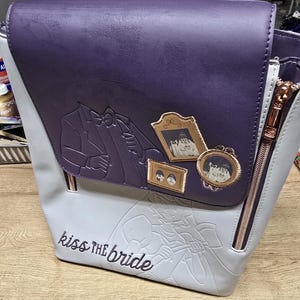 May include: A purple and gray backpack with a rose gold zipper. The flap features an embossed tuxedo design and three framed portraits. The phrase "kiss the bride" is embroidered on the front.