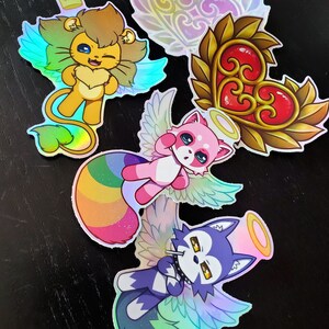 May include: Five iridescent holographic stickers featuring a lion, a panda, a wolf, and two heart designs. The lion has a crown and blue wings. The panda has pink fur and rainbow wings. The wolf has blue fur and rainbow wings. The hearts are gold with red centres.