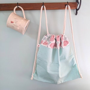 May include: A light blue drawstring bag with a floral print border. The bag is hanging on a wooden rack with white cords.