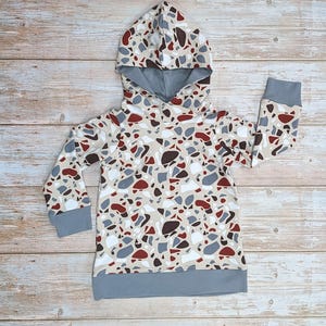 May include: A hooded sweatshirt for kids with a terrazzo pattern in shades of brown, red, gray, and white. The sweatshirt has a gray hood and a gray band at the bottom.