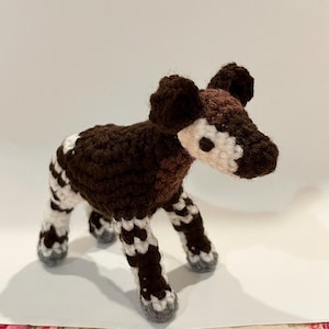 May include: A handmade crochet okapi figurine. The okapi has a brown body, a white and brown head, and white and black striped legs. The ears are brown, and the hooves are gray. The okapi is standing on a white surface.