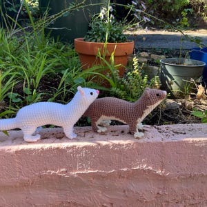 May include: Two crocheted ferret toys standing on a ledge. One ferret is white with a black-tipped tail and a pink nose, while the other is light brown with a cream-colored underbelly and face. Both have small dark eyes and stitched details.