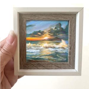 May include: A framed painting of a seascape with a sunset over the ocean. The painting features a blue sky with clouds, a golden sunset, and white waves crashing on the shore.