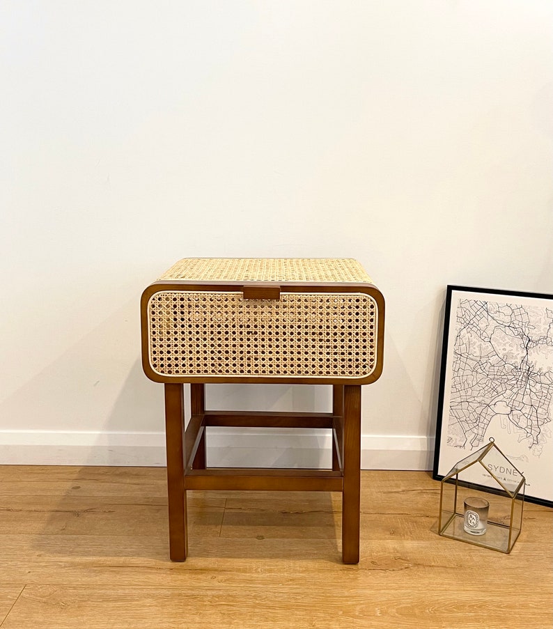 Op de afbeelding: Een kleine houten bijzettafel met een geweven rotan lade en een lager plank. De tafel heeft een donkerbruine afwerking en staat op vier poten.