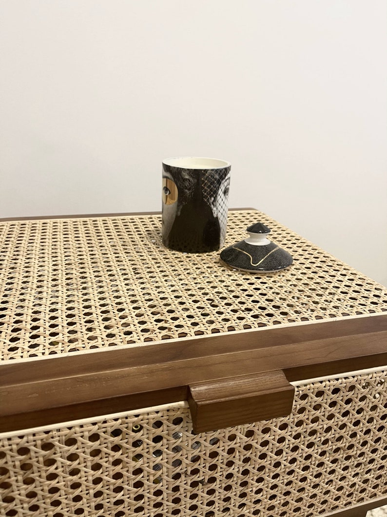 Op de afbeelding: Een bruine houten doos met een geweven rotan deksel. De doos heeft een bruine houten handgreep. Een zwart-witte keramische mok met een vrouwengezichtsontwerp en een zwart-witte keramische deksel met een gouden accent staan op de doos.