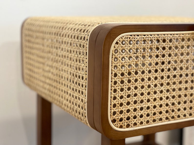 Op de afbeelding: Een close-up van een houten bijzettafel met een gevlochten rieten blad. De tafel heeft een lichtbruin houten frame en een witte rand rond het rieten blad.