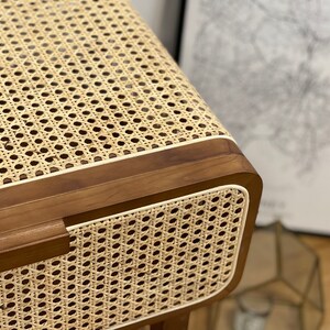 Op de afbeelding: Close-up van een houten bijzettafel met een geweven rotan blad en een witte rand. De tafel heeft een lade met een houten handvat.