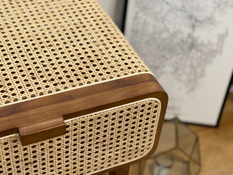 Op de afbeelding: Close-up van een houten bijzettafel met een geweven rotan blad en een witte rand. De tafel heeft een lade met een houten handvat.