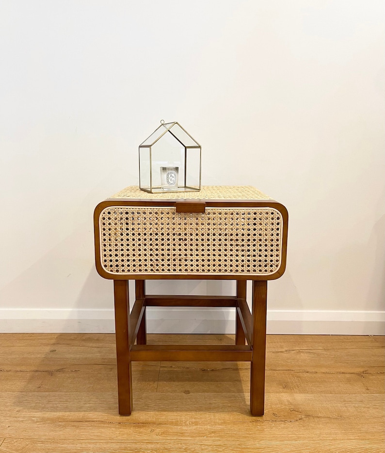 Op de afbeelding: Een klein houten nachtkastje met een geweven rotan lade en een lager plankje. Het nachtkastje heeft een lichtbruine afwerking en is versierd met messing beslag. Een klein glazen huis met een kaars erin staat op het nachtkastje.