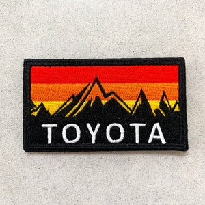 May include: Black and white embroidered patch with a sunset mountain scene and the word "TOYOTA" in white lettering.