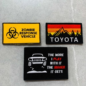 Könnte beinhalten: Drei gestickte Patches mit schwarzen Nähten. Der erste Patch ist gelb mit einem Biohazard-Symbol und dem Text "Zombie Response Vehicle". Der zweite Patch ist schwarz mit einer Bergkette und dem Text "Toyota". Der dritte Patch ist schwarz mit einer weißen Silhouette eines Jeeps und dem Text "The more I play with it the bigger it gets".