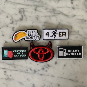 Puede incluir: Cinco parches bordados para planchar con diferentes diseños. Los parches incluyen un taco con el texto "Let's Taco Bout It", una persona corriendo con el texto "4-3-ER", un diablo rojo y negro con un logotipo de Toyota, una bolsa de la compra con el texto "Certified Mall Crawler", y una bomba de gasolina con el texto "Heavy Drinker".