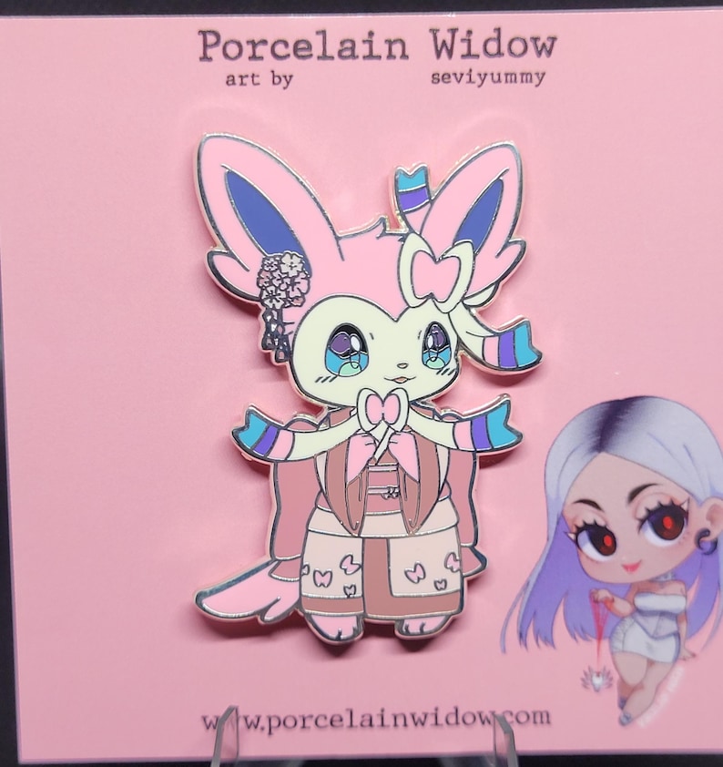 May include: A pink enamel pin featuring a Sylveon-inspired character in a kimono. The pin has a bow, floral accents, and the text "Porcelain Widow" and "seviyummy". The character has large, expressive eyes.