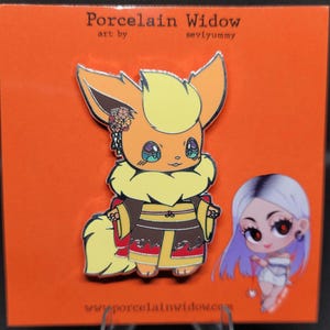 May include: A colorful enamel pin featuring an orange and yellow cartoon character with a kimono. The pin is on an orange backing card with the text "Porcelain Widow" and "seviyummy". A second cartoon character is also on the card.