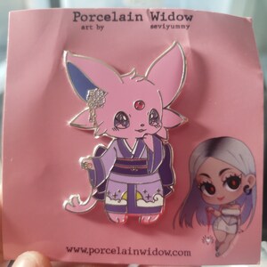May include: A pink enamel pin featuring a cartoon fox-like character in a kimono. The pin has a silver outline and includes the text "Porcelain Widow" and "seviyummy". The character has a red gem on its forehead and is holding its chin.