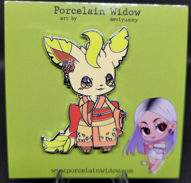 May include: A pin featuring a cartoon fox-like character in a yellow and orange kimono. The pin has a floral hair accessory and is set against a green background. The text "Porcelain Widow" and "seviyummy" are visible.