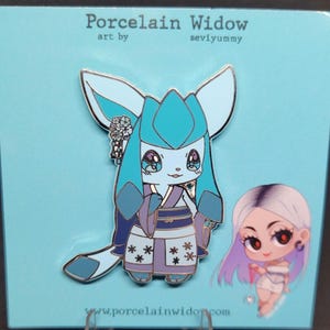 May include: A blue enamel pin featuring a cartoon character with large ears and a kimono. The pin has a light blue and purple color scheme, with the text "Porcelain Widow" at the top. A smaller pin of a girl with purple hair is also visible.
