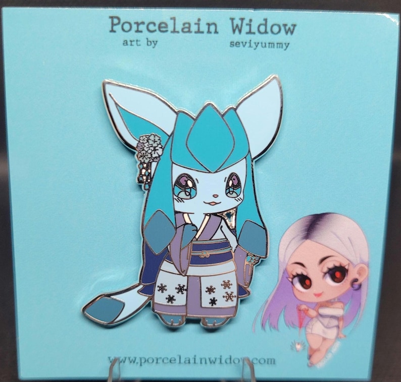 May include: A blue enamel pin featuring a cartoon character with large ears and a kimono. The pin has a light blue and purple color scheme, with the text "Porcelain Widow" at the top. A smaller pin of a girl with purple hair is also visible.
