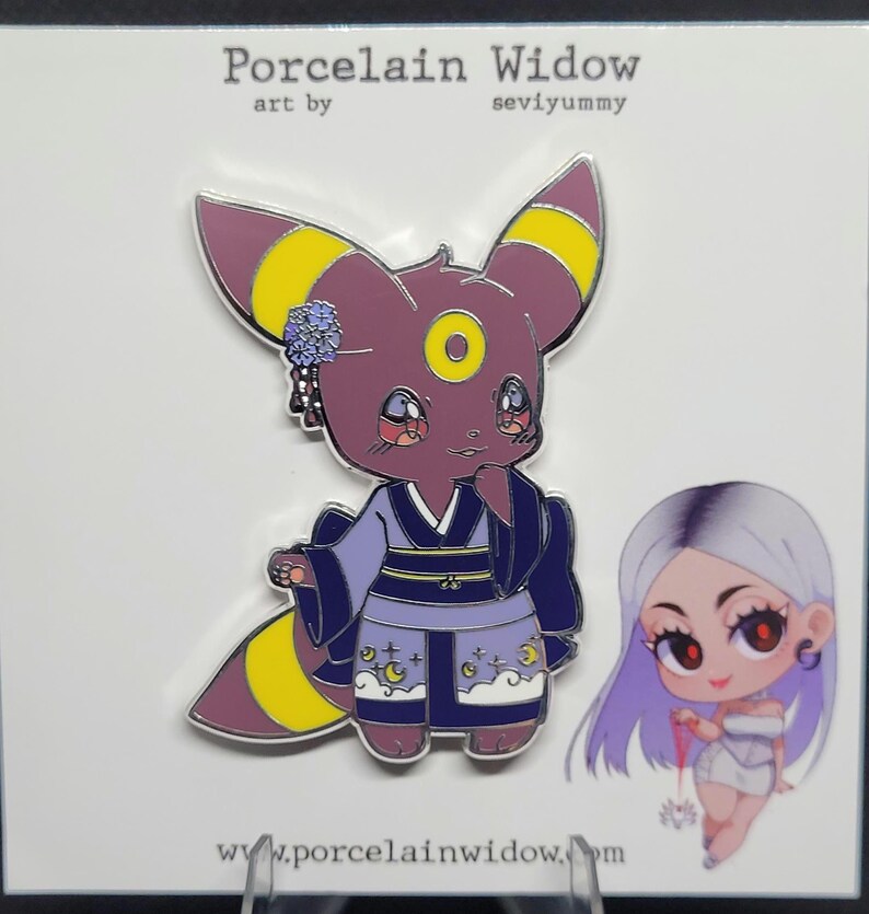 May include: A pin featuring a cartoon Umbreon character in a kimono with a moon and star design. The pin has purple, yellow, and blue accents. The text "Porcelain Widow" and "seviyummy" are at the top, and "www.porcelainwidow.com" is at the bottom.