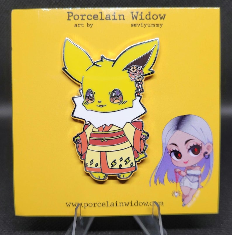 May include: A yellow enamel pin featuring a cartoon fox-like character in a kimono. The pin has a white collar and red and yellow kimono. The pin is on a yellow backing card with the text "Porcelain Widow" and "seviyummy".