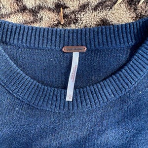 Puede incluir: Un su&eacute;ter azul oscuro con una etiqueta de Free People. La etiqueta est&aacute; unida al su&eacute;ter con un peque&ntilde;o alfiler. El su&eacute;ter est&aacute; hecho de una tela suave y de punto.