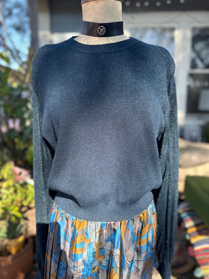 Puede incluir: Un su&eacute;ter azul oscuro con brillo, mangas largas y cuello redondo. El su&eacute;ter est&aacute; en un maniqu&iacute; y se combina con una falda estampada de colores.