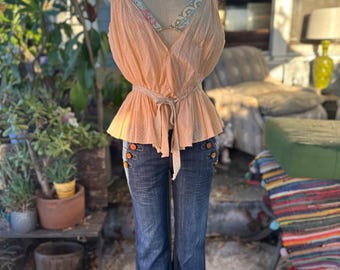 May include: A peach-colored, sleeveless top with a tie at the waist and sailor buttons. The top is worn over a floral patterned bra and paired with dark blue, wide-leg jeans.