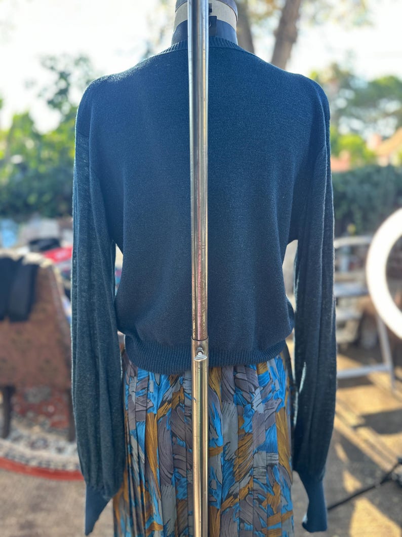 Puede incluir: Un su&eacute;ter azul teal de manga larga con dobladillo y pu&ntilde;os de canal&eacute;. El su&eacute;ter est&aacute; en un maniqu&iacute; y se combina con una falda estampada azul, dorada y blanca.