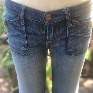 Puede incluir: Un par de pantalones vaqueros azules con cierre de botón y un lavado claro. Los vaqueros tienen un estilo de corte de bota y están hechos de una tela suave y cómoda.