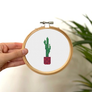 May include: A green cactus plant in a red pot, cross-stitched on white fabric in a wooden embroidery hoop.