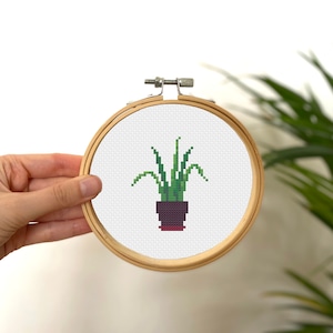 May include: A cross-stitch embroidery of a potted plant with green leaves in a dark purple pot with a red rim. The embroidery is framed in a wooden hoop.