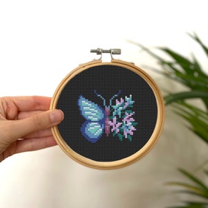 Könnte beinhalten: Ein blauer Schmetterling mit lila und rosa Blumen, gestickt auf einem schwarzen Stoff in einem hölzernen Stickrahmen.