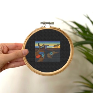 May include: A cross-stitch embroidery hoop with a black background and a colorful pixelated image of The Persistence of Memory by Salvador Dali.