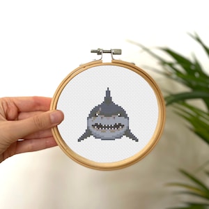 May include: A cross-stitch embroidery of a shark in a wooden hoop. The shark is depicted in shades of gray and white, with sharp teeth and a menacing expression. The wooden hoop is round and has a metal tightening mechanism.
