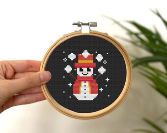 May include: A black and white cross-stitch pattern of a snowman wearing a red hat and scarf, holding snowballs in each hand. The snowman is surrounded by white snowflakes on a black background.