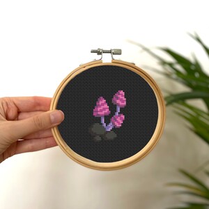 Könnte beinhalten: Kreuzstich-Stickerei mit drei rosa Pilzen mit lila Stielen und dunkelgrauen Kappen auf schwarzem Hintergrund. Das Design ist in einem runden Holz-Stickrahmen eingefasst. Das Kunstwerk wird von einer Hand gehalten.