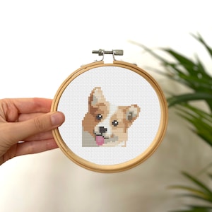 Puede incluir: Una bordado de punto de cruz de un perro corgi con un pecho blanco y un pelaje marrón y blanco. El perro está sacando la lengua y tiene una expresión feliz. El bordado está enmarcado en un aro de madera.