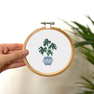 May include: A cross-stitch embroidery hoop with a green plant in a gray pot. The plant has large leaves and a long stem.