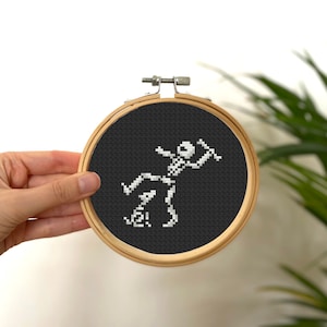 May include: A white skeleton cross-stitched on black fabric in a wooden embroidery hoop. The skeleton is dancing with the text "Yee!" below it.