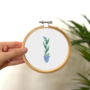 Puede incluir: Una bordado de punto de cruz de una planta en maceta con flores azules y hojas verdes en un aro de bordado de madera.