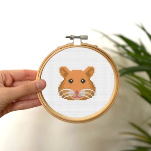 May include: A cross-stitch embroidery of a hamster's face in a wooden hoop. The hamster is rendered in shades of brown, with black eyes and pink details. The hoop is held in a hand, with a white background.