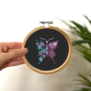Könnte beinhalten: Ein lilafarbener Schmetterling mit floralen Details, gestickt auf schwarzem Stoff in einem hölzernen Stickrahmen.