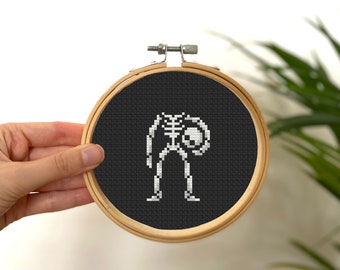 May include: A black and white cross-stitch embroidery hoop with a skeleton holding a skull. The skeleton is white and the background is black.