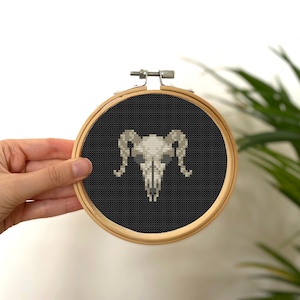 May include: A black and white cross-stitch embroidery of a ram skull with horns, framed in a wooden hoop.