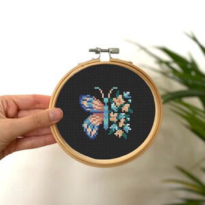 Könnte beinhalten: Ein Kreuzstich-Stickrahmen mit schwarzem Hintergrund, auf dem ein blauer und rosa Schmetterling mit einem Blumenmuster abgebildet ist.