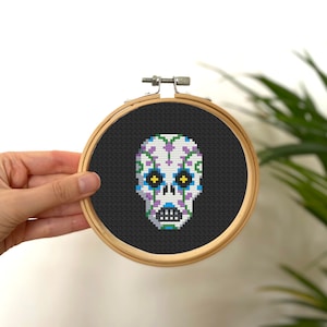 May include: A white sugar skull with purple and blue floral designs and yellow eyes is embroidered on black fabric in a wooden embroidery hoop.