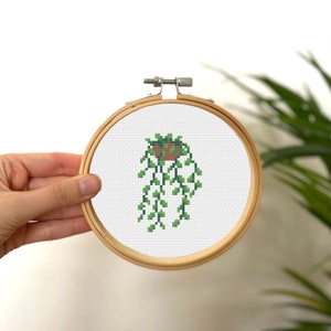 May include: A cross-stitch embroidery of a hanging plant in a brown pot with green leaves. The embroidery is framed in a wooden hoop.