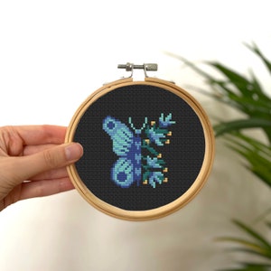 Könnte beinhalten: Kreuzstich-Stickerei eines Schmetterlings und eines floralen Designs in einem Holzreifen. Der Schmetterling ist in Blautönen gehalten und der Hintergrund ist schwarz. Der Reifen wird von einer Hand gehalten.