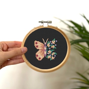 Könnte beinhalten: Kreuzstich-Stickerei eines Schmetterlings und Blumen in einem Holzreifen. Der Schmetterling ist hellrosa mit dunkelrosa Punkten, und die Blumen sind grün und lila auf schwarzem Hintergrund. Der Reifen wird von einer Hand gehalten.