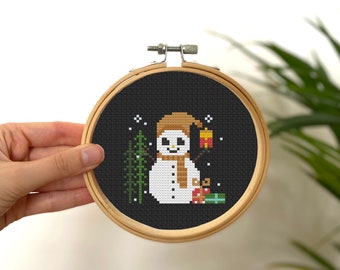May include: A black and white cross-stitch pattern of a snowman holding a gift and standing next to a small evergreen tree. The snowman is wearing a brown hat and a tan scarf. The cross-stitch is framed in a round wooden hoop.